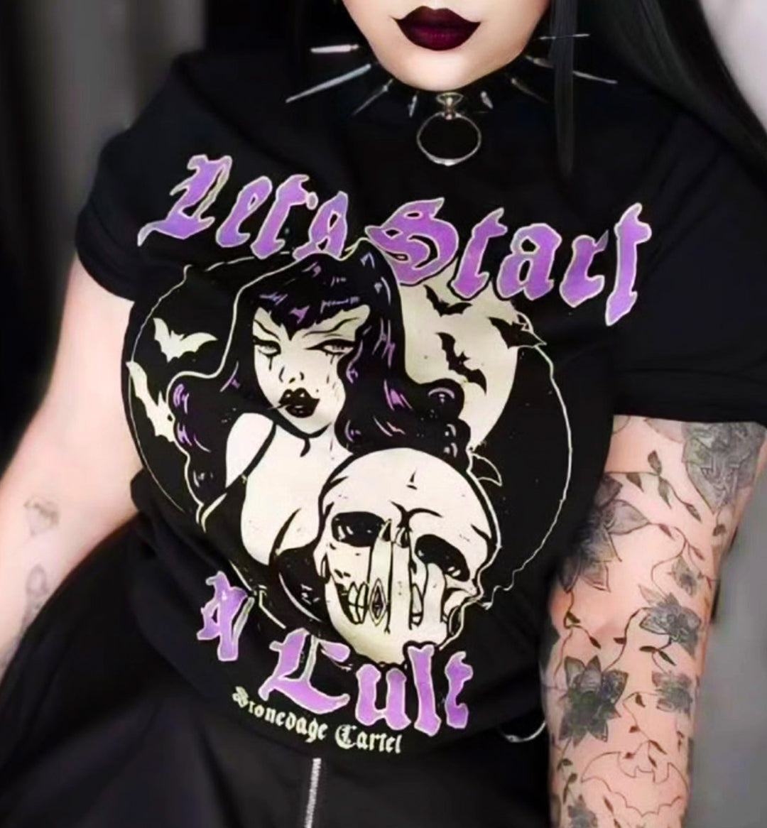 A close-up of a goth girl wearing a black graphic T-shirt with a gothic design featuring a woman's face, a skull, bats, and purple text reading 'Let's Start a Cult' and 'Stonedage Cartel'. The person has dark hair, black lipstick, and visible arm tattoos.