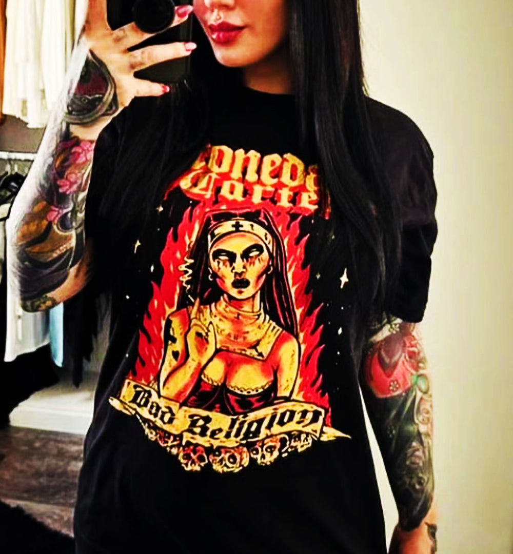 A person taking a mirror selfie wearing a black t-shirt with a large graphic print. The graphic features a gothic-style illustration of a woman in a nun's habit with the text 'Stonedage Cartel' and 'Bad Religion' in a red, yellow, and black design.