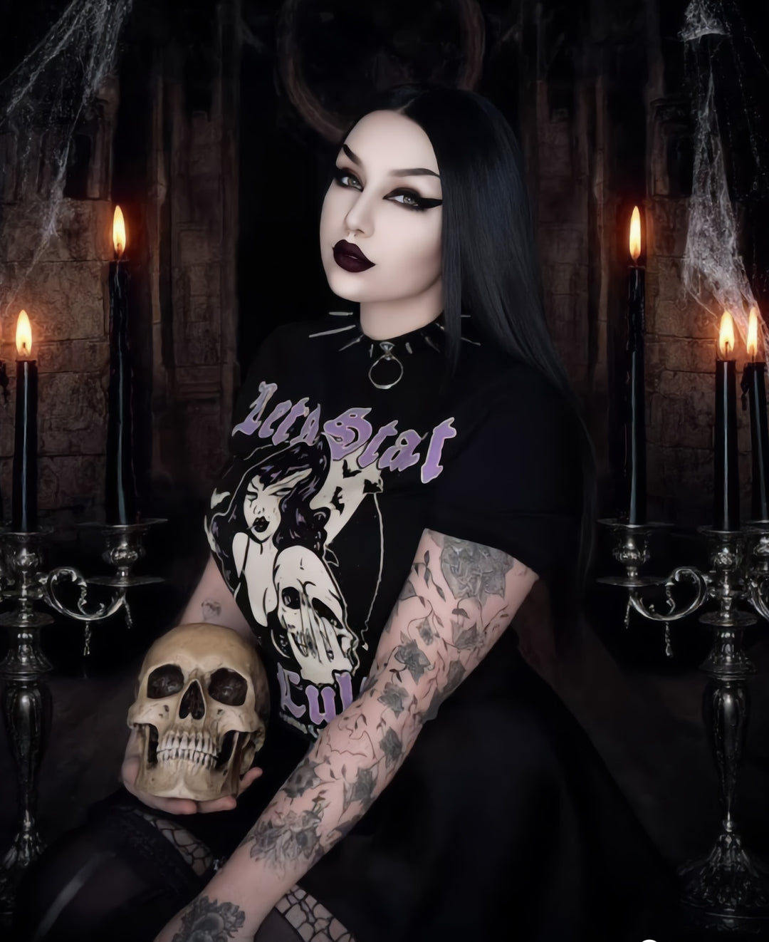 Person wearing a black t-shirt with graphic design and text, holding a skull in a dark, candle-lit setting.