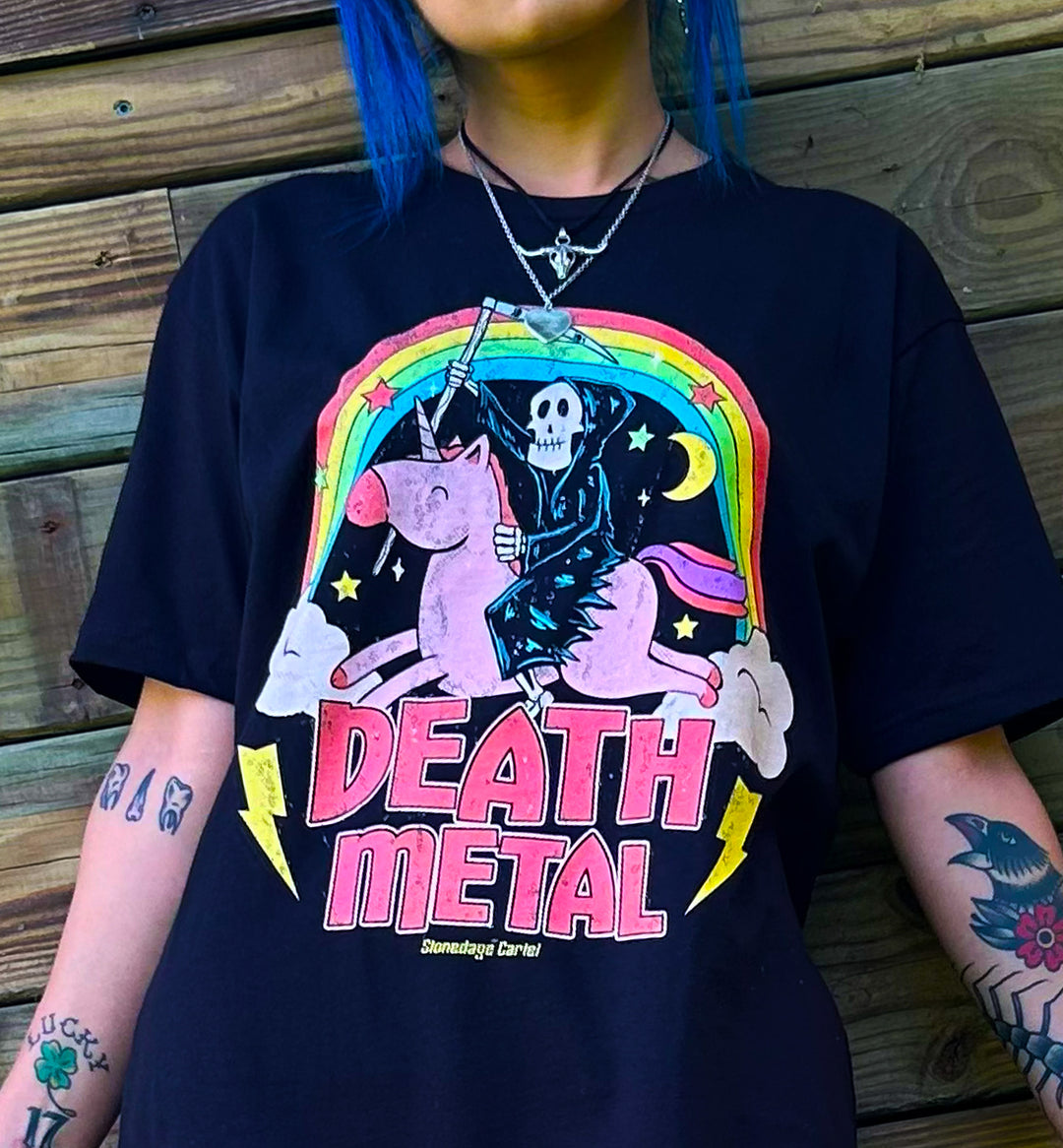 A person wearing a black graphic t-shirt with a colorful 'Death Metal' band design featuring a skeleton riding a pink unicorn under a rainbow. The person has blue hair and visible arm tattoos, standing against a rustic wooden wall.