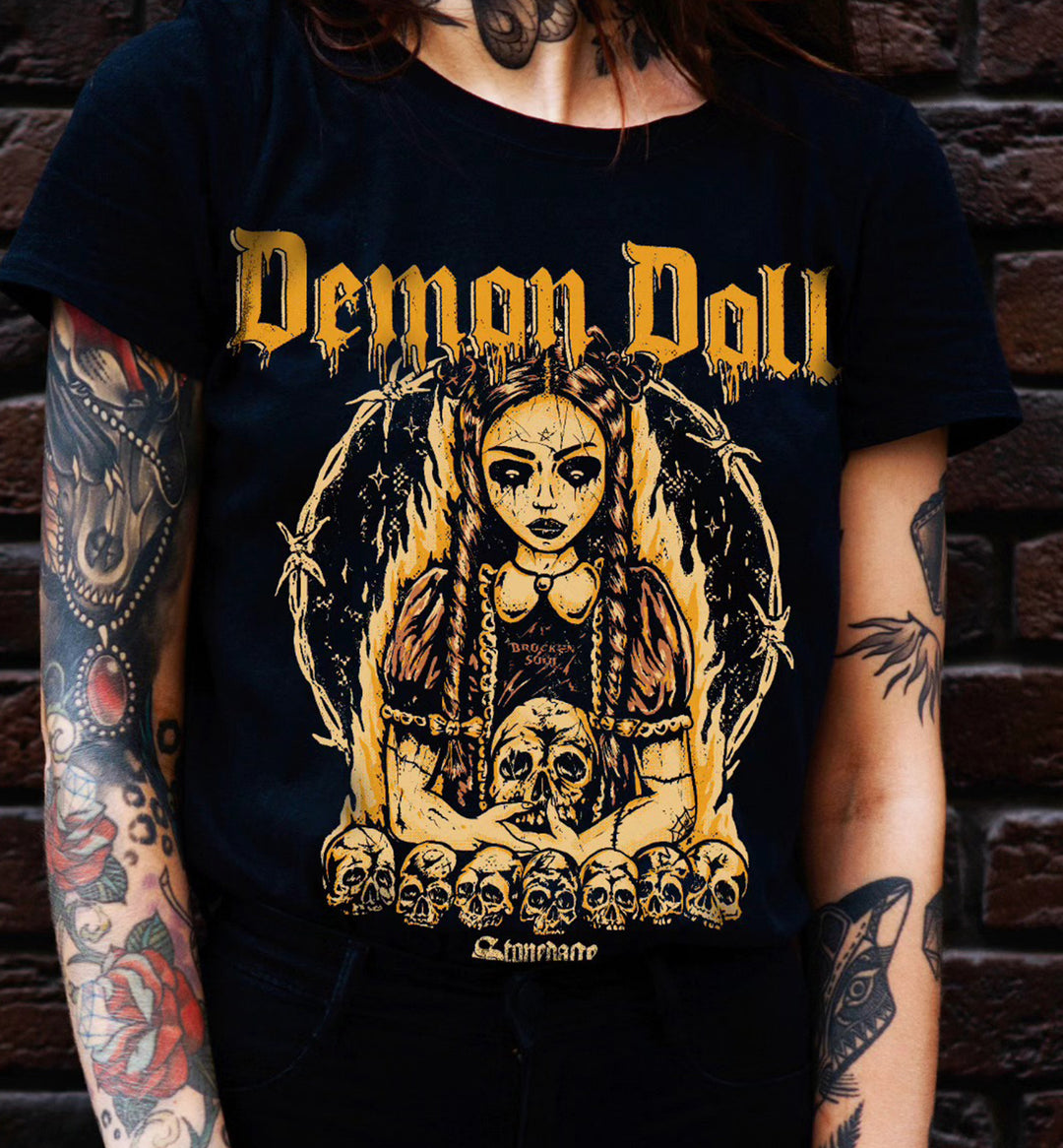 A person wearing a black graphic T-shirt with a large yellow and gold illustration and the text 'Demon Doll' on the front, standing against a dark brick wall.