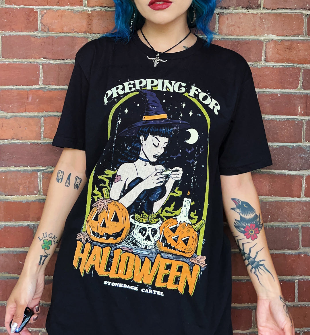 A person wearing a black graphic T-shirt with a colorful Halloween-themed illustration and the text 'PREPPING FOR HALLOWEEN' and 'STONEDAGE CARTEL', standing against a red brick wall.
