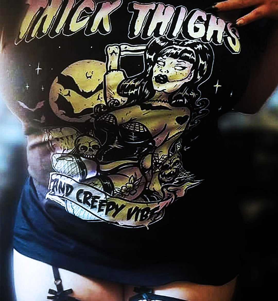 A close-up of a person wearing a black graphic t-shirt with a creepy vintage-style illustration and the text 'THICK THIGHS' and 'AND CREEPY VIBE'.